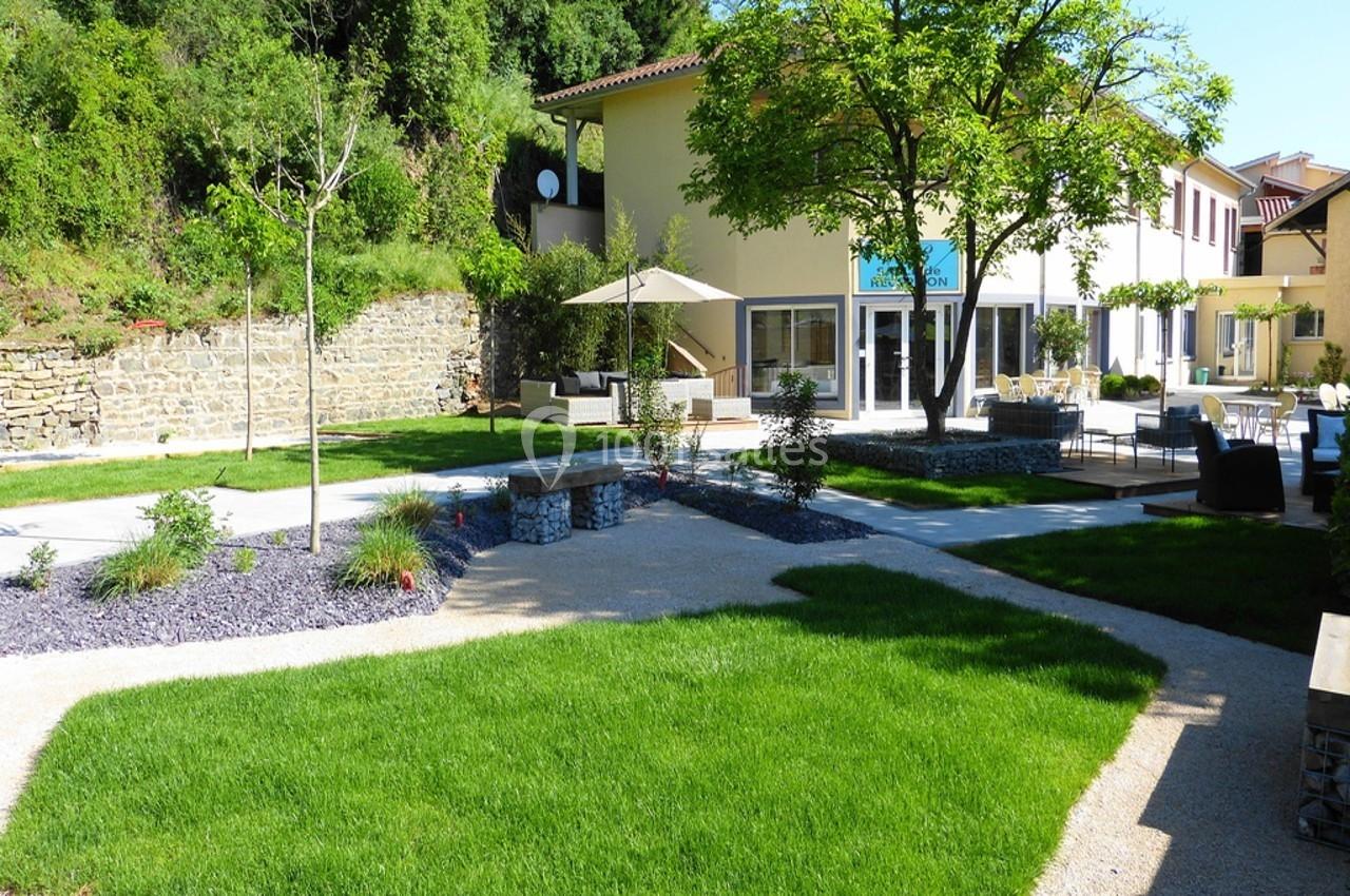 Jardin aménagé avec pelouse, allées gravillonnées, bancs et arbres, entouré de bâtiments modernes et végétation.