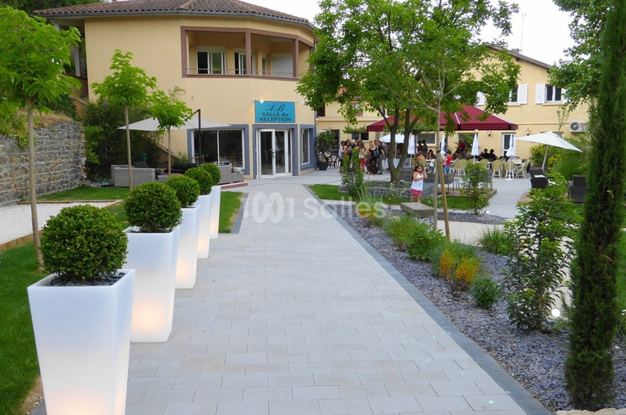 Allée pavée bordée de pots lumineux menant à un bâtiment avec terrasse et espaces verts aménagés.