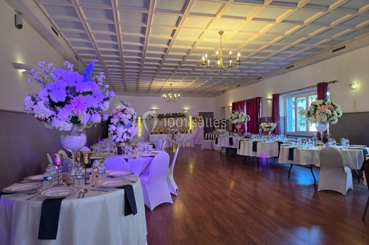 Salle de réception décorée avec des tables dressées, des fleurs blanches et violettes, et un éclairage chaleureux.