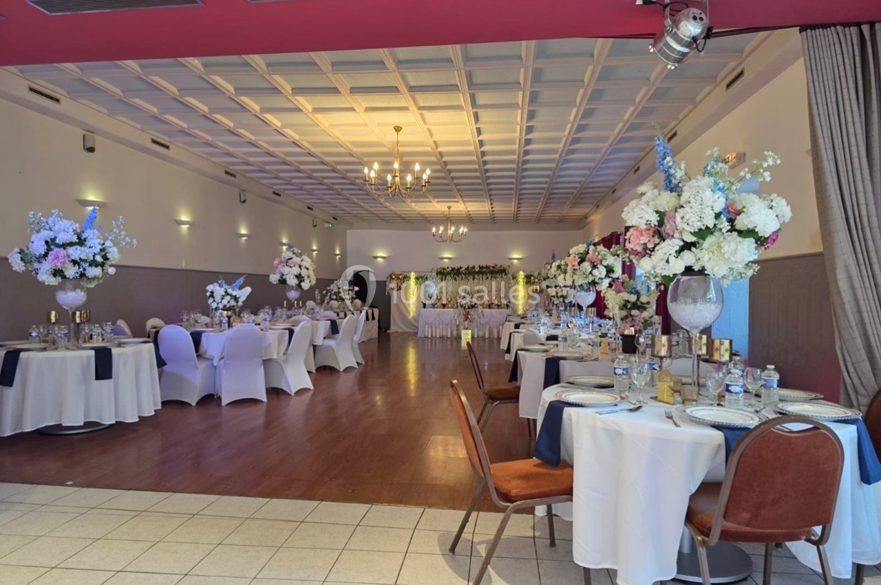 Salle de réception décorée pour un événement, avec tables dressées, chaises couvertes et arrangements floraux.