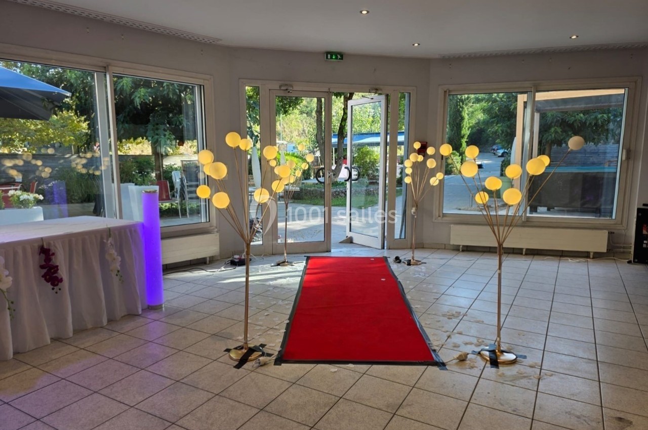 Entrée d'une salle avec tapis rouge, décorée de lampes sphériques et donnant sur un espace extérieur arboré.
