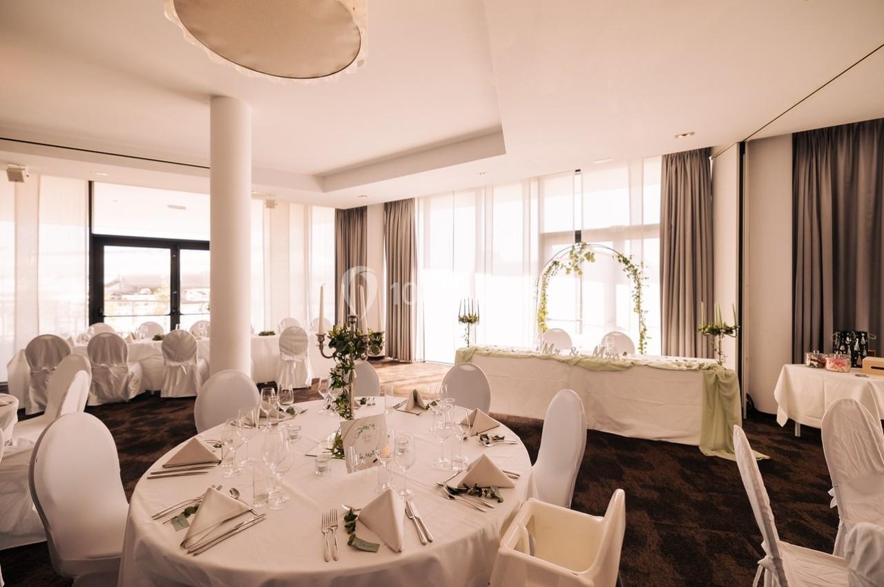 Salle de réception lumineuse avec tables rondes dressées, chaises blanches et une arche décorée de fleurs.