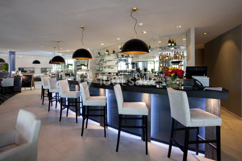 Location salle Mons (Hainaut) - Congrès Hôtel Mons Van Der Valk #2 Bar moderne avec comptoir éclairé, tabourets rembourrés, suspensions noires et étagères garnies de bouteilles.