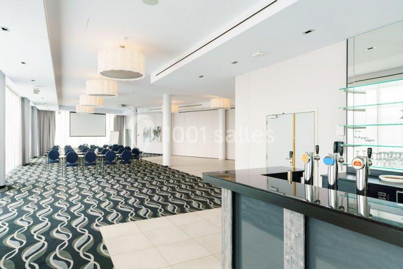 Location salle Mons (Hainaut) - Congrès Hôtel Mons Van Der Valk #5 Salle de conférence lumineuse avec rangées de chaises, écran de projection et bar équipé au premier plan.