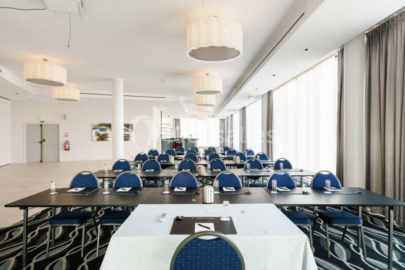 Location salle Mons (Hainaut) - Congrès Hôtel Mons Van Der Valk #4 Salle de réunion lumineuse avec des tables noires, des chaises bleues et des rideaux gris encadrant de grandes fenêtres.