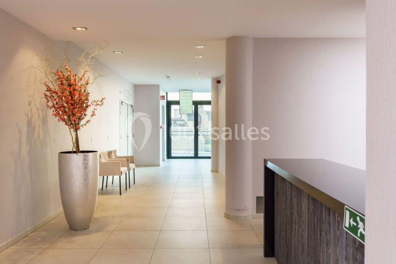 Location salle Mons (Hainaut) - Congrès Hôtel Mons Van Der Valk #7 Un hall lumineux avec un comptoir sombre, un grand vase de fleurs orange et des chaises près d'une porte vitrée.