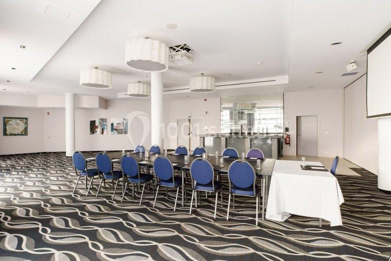 Location salle Mons (Hainaut) - Congrès Hôtel Mons Van Der Valk #10 Salle de réunion lumineuse avec des chaises bleues autour de tables noires, un bar en arrière-plan et un écran blanc.