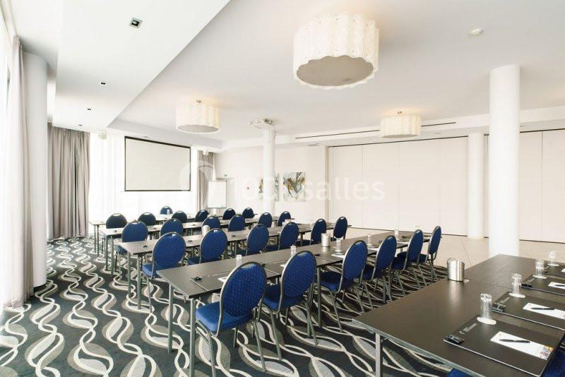 Location salle Mons (Hainaut) - Congrès Hôtel Mons Van Der Valk #11 Salle de conférence lumineuse avec tables noires, chaises bleues, écran de projection et décor moderne.