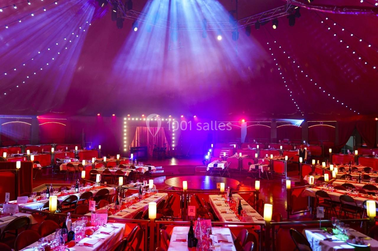 Salle de spectacle sous un chapiteau avec tables dressées, éclairages colorés et scène illuminée en arrière-plan.