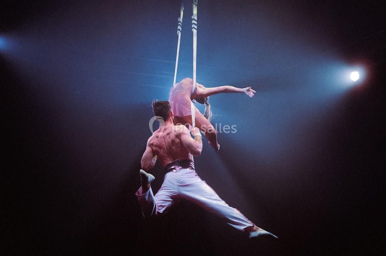 Deux artistes de cirque exécutent une figure acrobatique suspendus à des sangles sous un éclairage de scène.