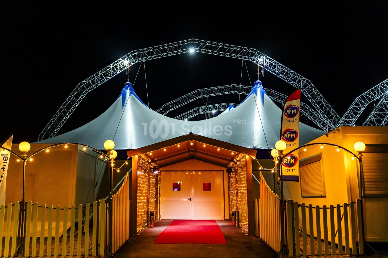 Entrée d’un chapiteau illuminé la nuit, avec tapis rouge, guirlandes lumineuses et structures métalliques visibles.