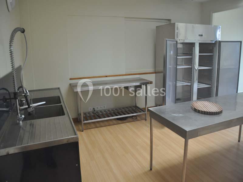 Cuisine professionnelle avec évier, tables en inox, réfrigérateur ouvert et plan de travail en bois sur sol clair.