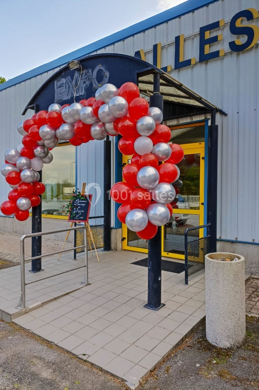Entrée d'un bâtiment décorée d'une arche de ballons rouges et argentés, avec une rampe d'accès et une ardoise.
