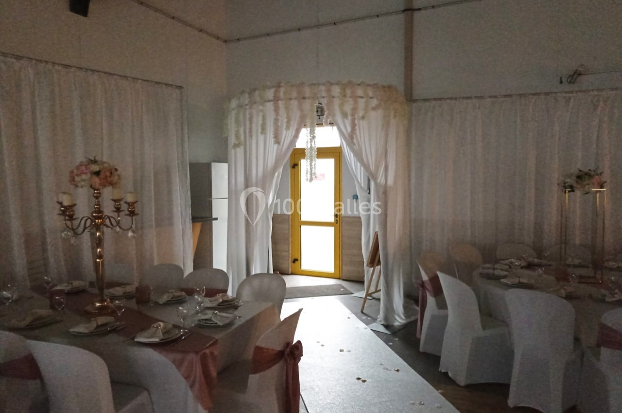 Salle de réception décorée avec des chaises blanches, des nappes roses et une arche ornée de fleurs près d'une porte.