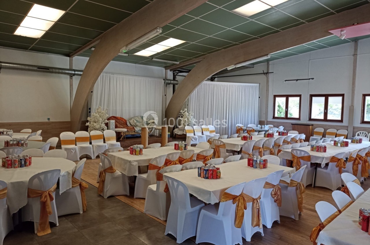 Salle décorée pour un événement, avec des tables rondes dressées, nappes blanches et chaises ornées de nœuds orange.