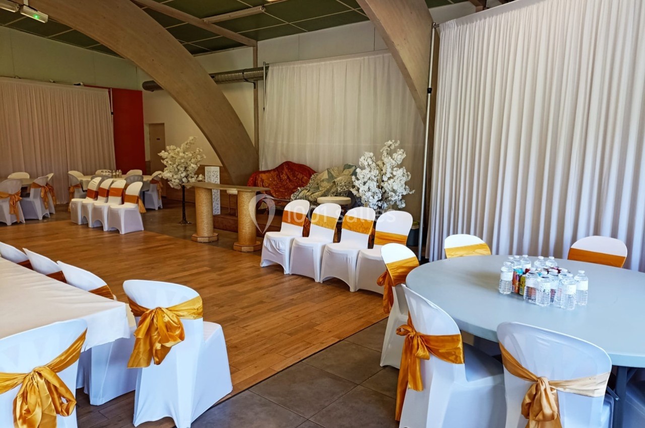 Salle décorée pour un événement avec des chaises blanches ornées de nœuds dorés, tables dressées et rideaux blancs.