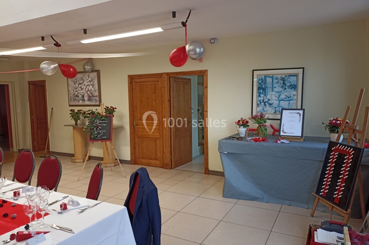 Salle décorée pour un événement avec tables dressées, ballons rouges, ardoise et table de présentation ornée de fleurs.