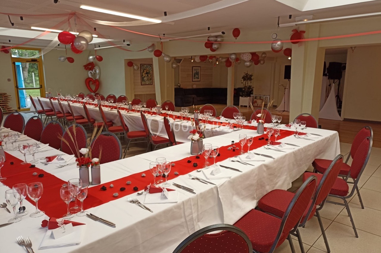 Salle décorée pour un événement avec tables dressées en U, nappes blanches, chemins de table rouges et ballons suspendus.