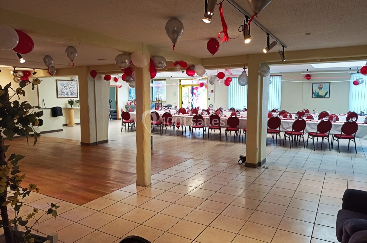 Salle de réception décorée avec des ballons rouges et blancs, tables dressées en rangées pour un événement.