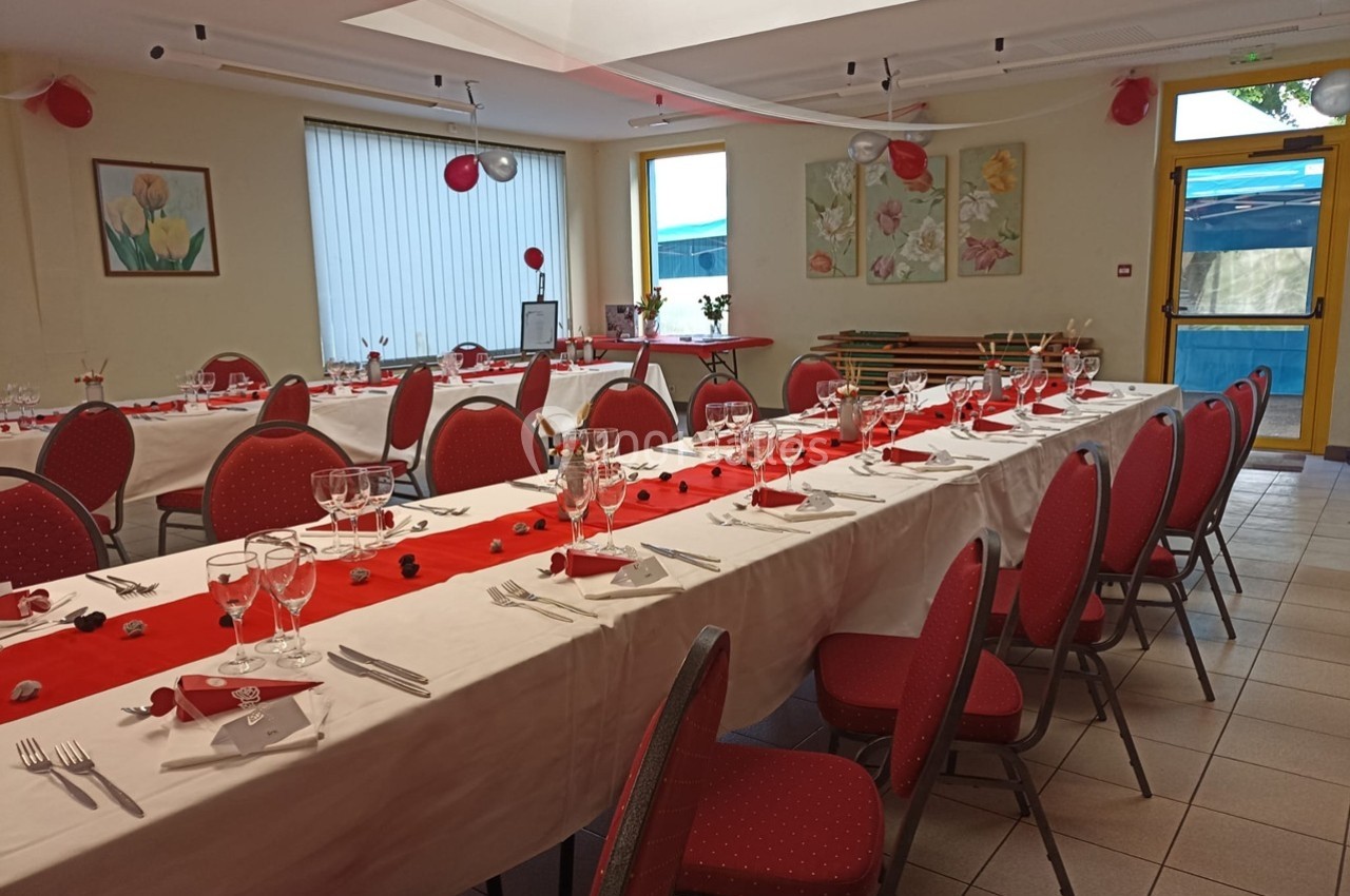 Salle décorée pour un événement, avec longues tables dressées, nappes blanches et chemins de table rouges.
