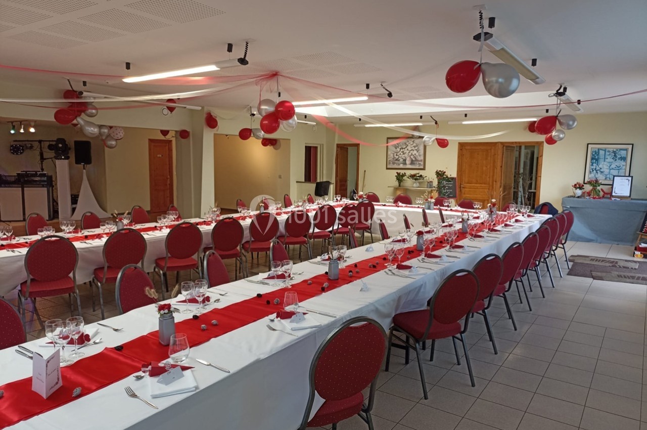 Salle décorée pour un événement, avec longues tables dressées, nappes blanches, chemins de table rouges et ballons suspendus.
