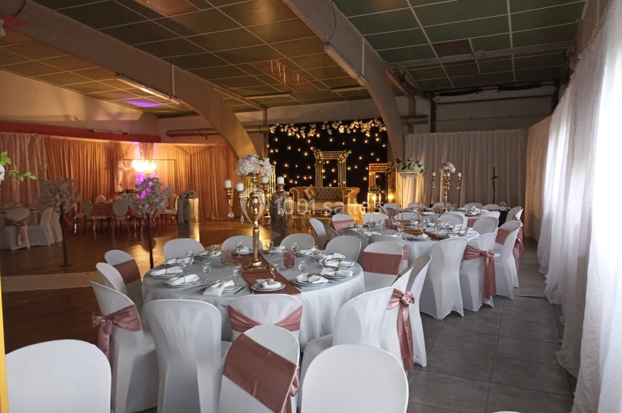 Salle de réception décorée avec des tables rondes dressées, des chaises blanches ornées de rubans roses et un espace…