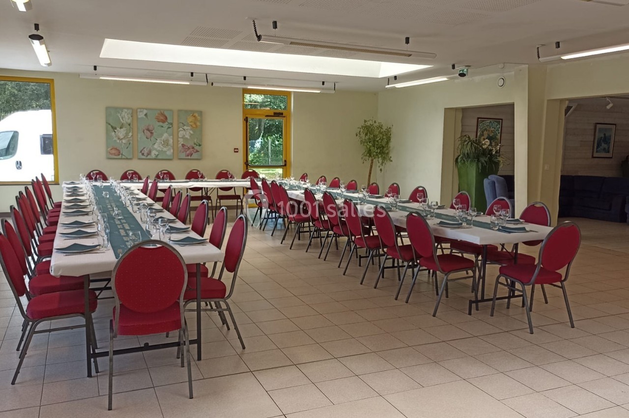 Salle lumineuse aménagée avec deux longues tables dressées pour un repas, entourées de chaises rouges.