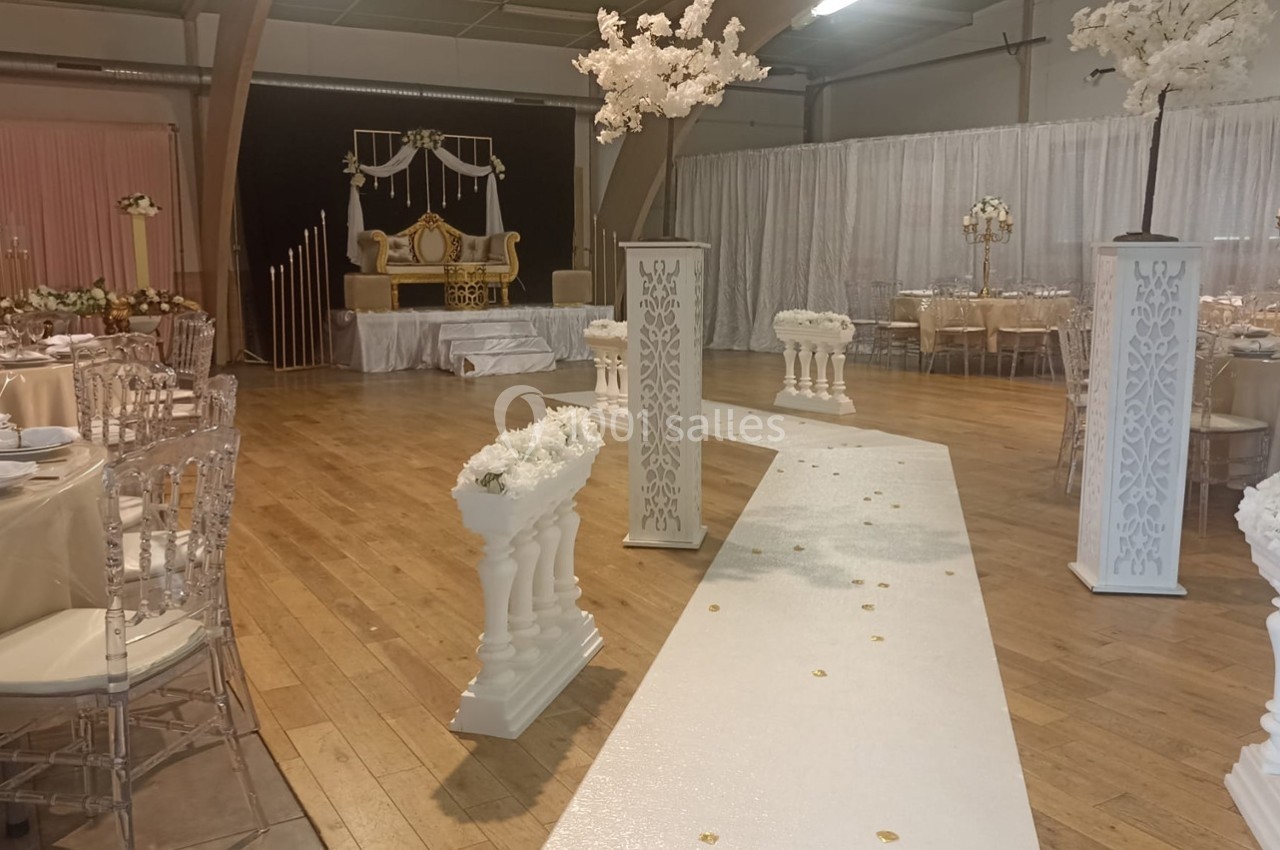 Salle décorée pour un mariage avec allée blanche ornée de fleurs, chaises transparentes et table d'honneur en arrière-plan.