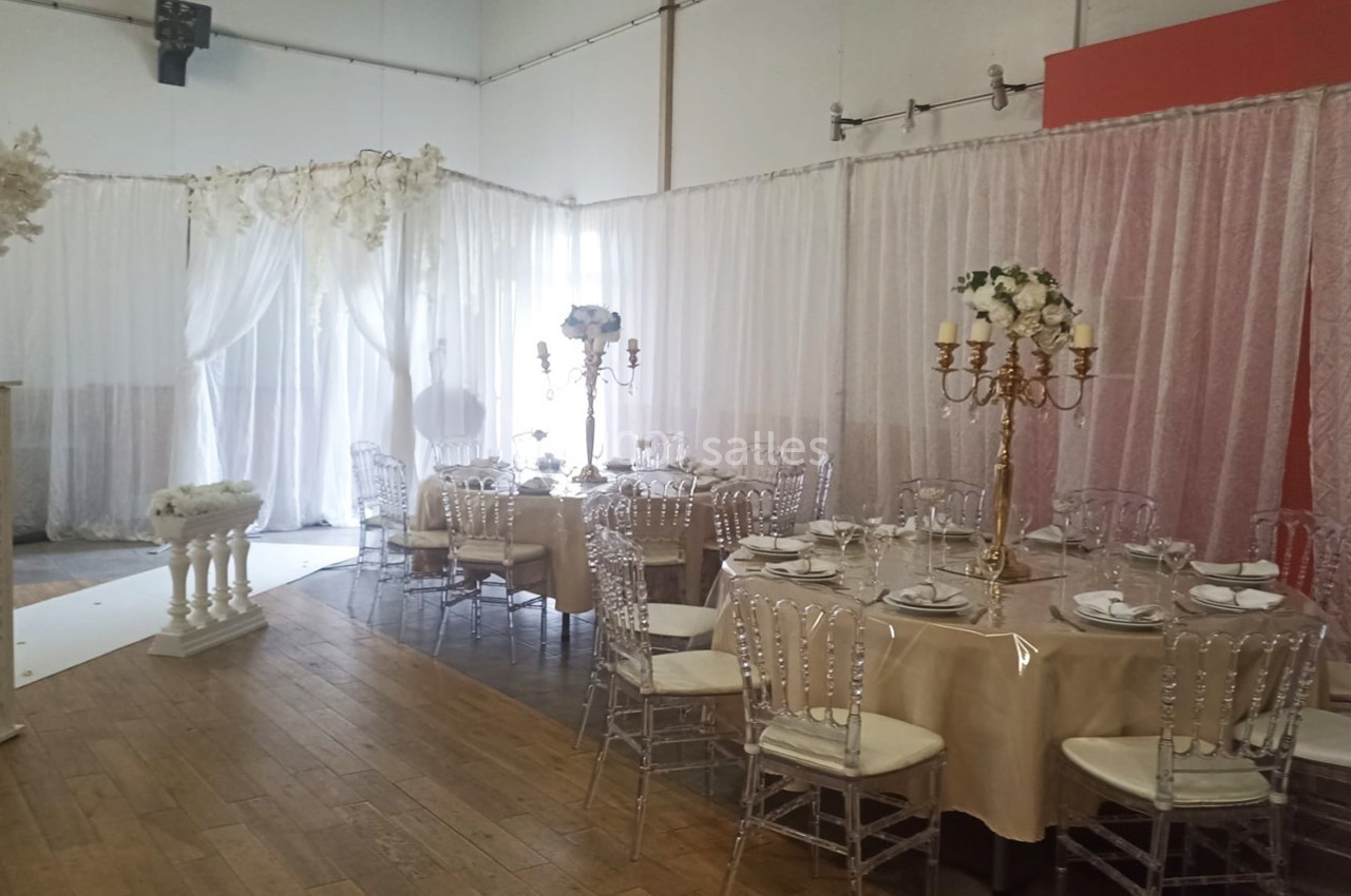 Salle décorée pour un événement avec tables rondes, nappes beige, chaises transparentes et rideaux blancs et roses.