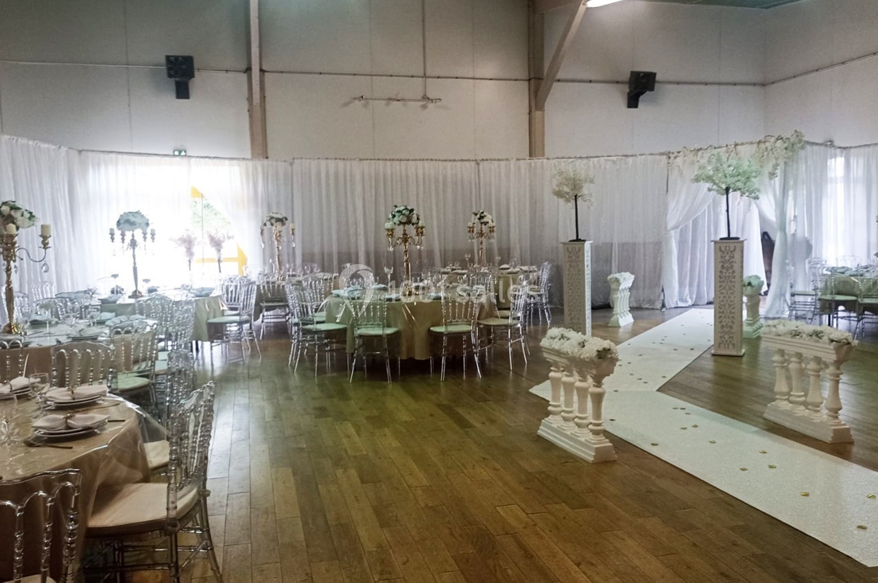 Salle de réception décorée avec des tables rondes, des chaises transparentes et une allée ornée de colonnes et fleurs…