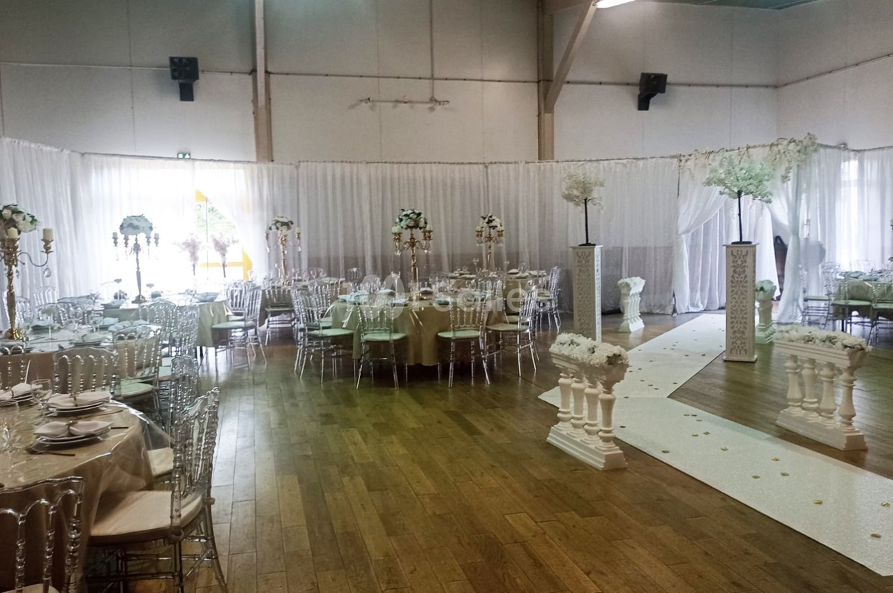 Salle de réception décorée avec des tables rondes, des chaises transparentes et une allée ornée de colonnes et fleurs…
