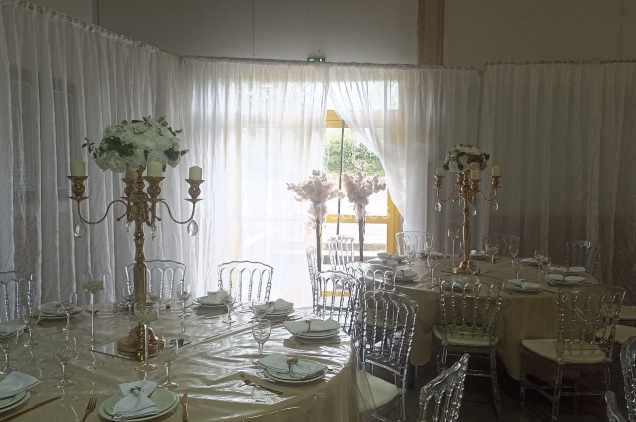 Salle décorée pour un événement avec tables rondes, nappes beiges, chaises transparentes et chandeliers dorés ornés de…