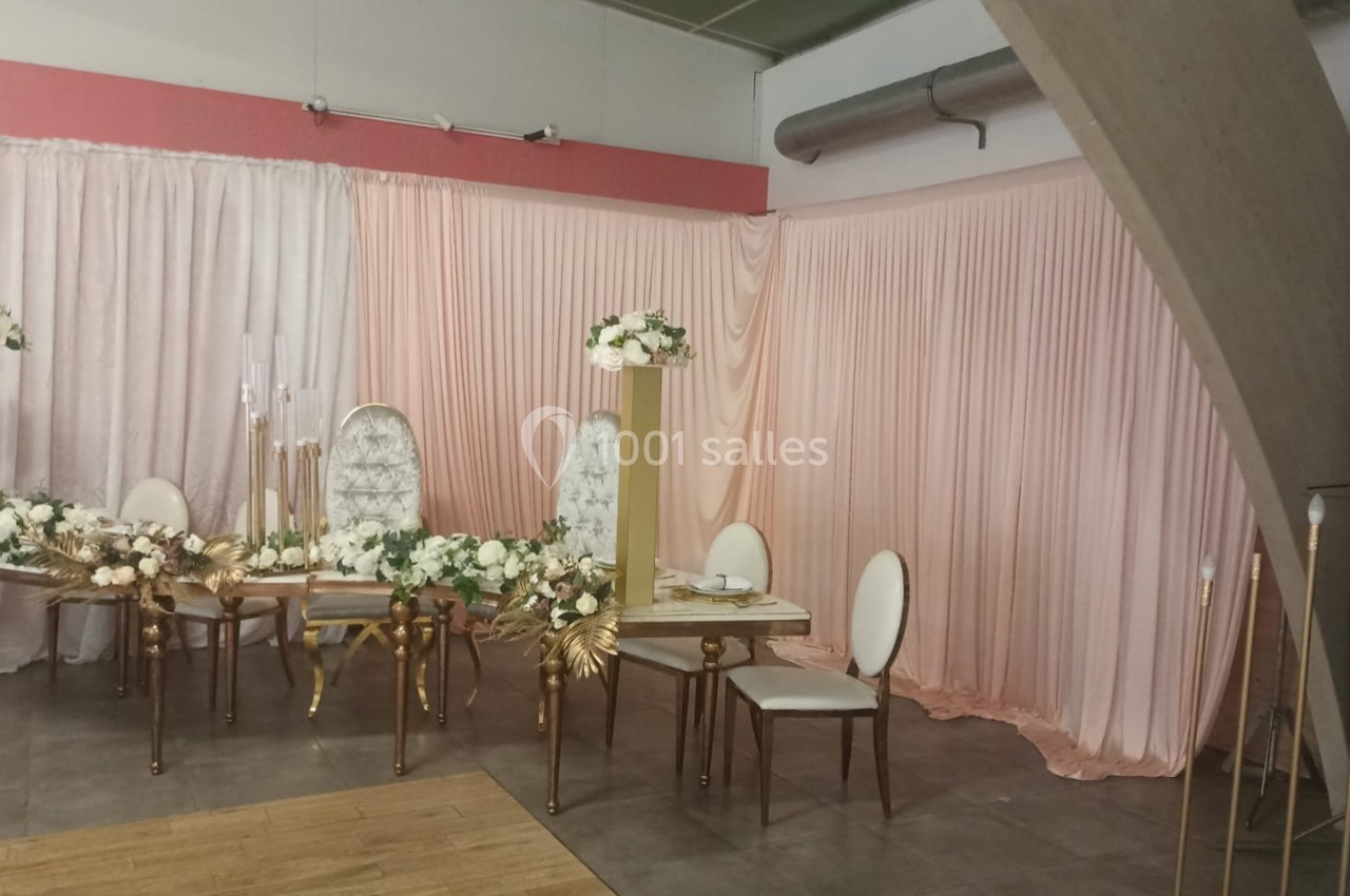 Salle décorée pour un événement avec des chaises blanches, une table dorée et des rideaux roses en arrière-plan.