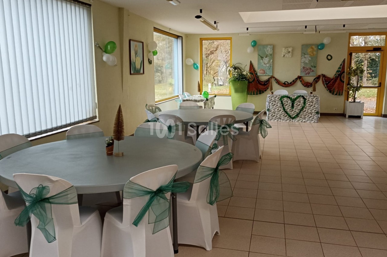 Salle décorée pour un événement, avec des tables rondes, des chaises ornées de rubans verts et une table d'honneur.
