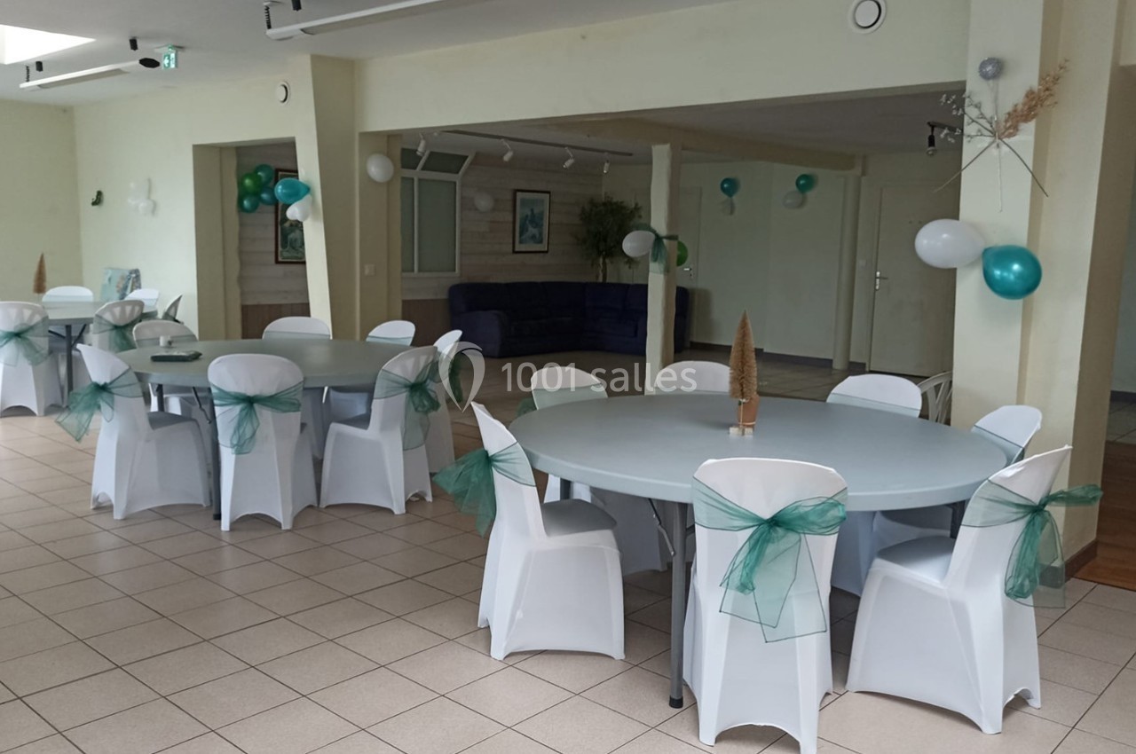 Salle décorée pour un événement avec des tables rondes, des chaises blanches ornées de rubans verts et des ballons.