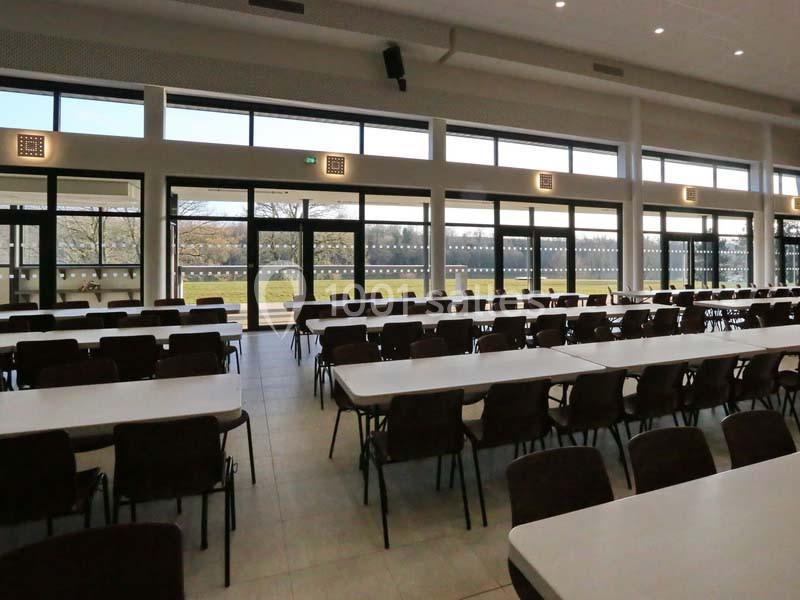 Salle spacieuse avec de grandes baies vitrées, des rangées de tables et chaises, donnant sur un espace extérieur verdoyant.