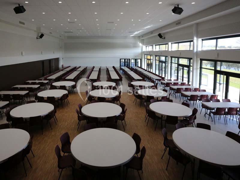 Salle spacieuse avec de grandes tables rondes et des chaises alignées, éclairée par des baies vitrées.