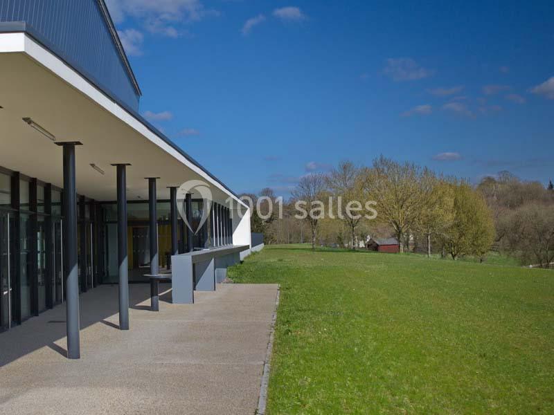 Bâtiment moderne avec une terrasse couverte, bordé d'une pelouse et d'arbres sous un ciel bleu.