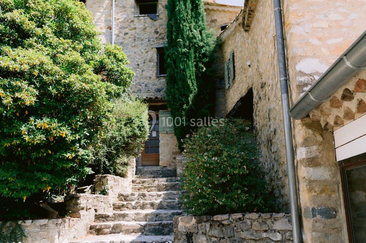 Escalier en pierre bordé de pots de fleurs menant à une maison en pierre avec un grand cyprès et des arbustes.