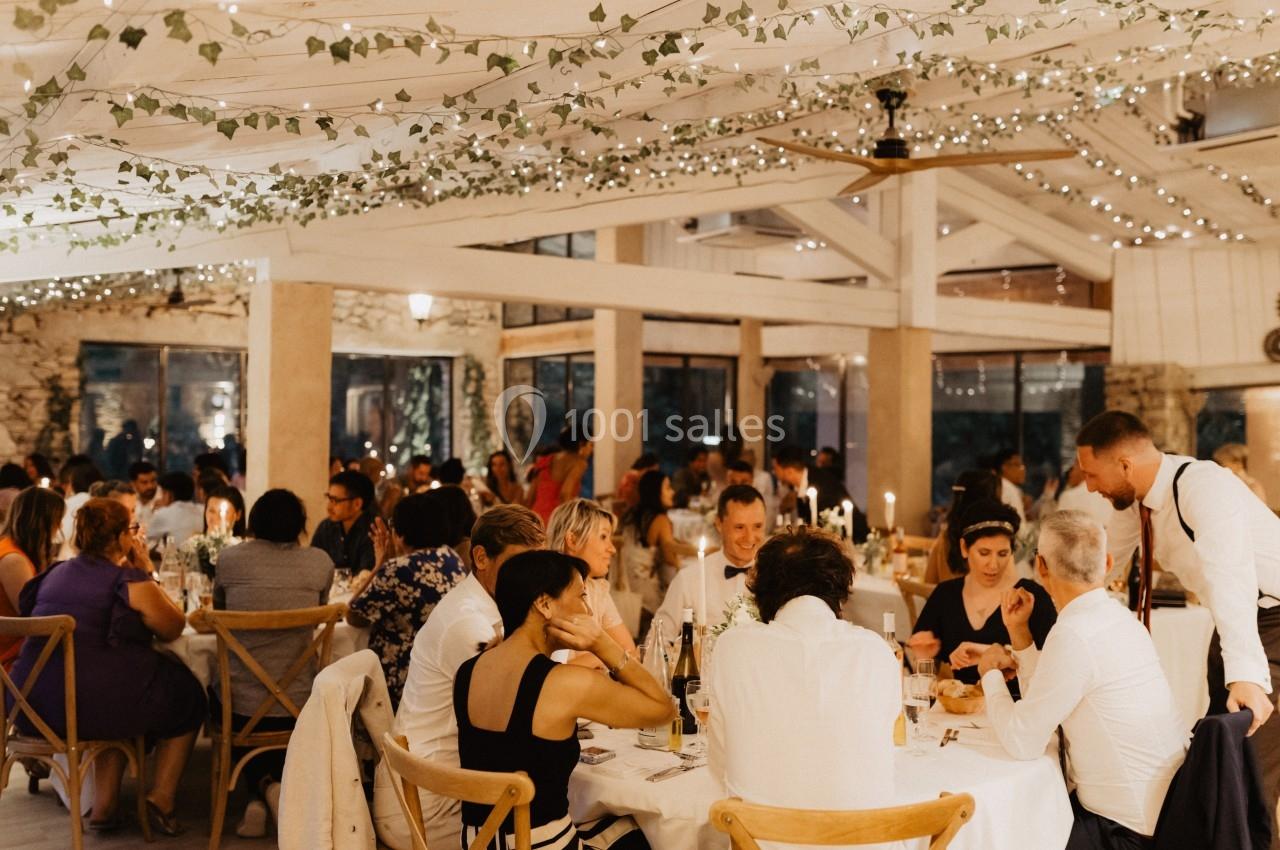 Salle de réception lumineuse avec des invités dînant à des tables décorées, sous des guirlandes lumineuses et des plantes…