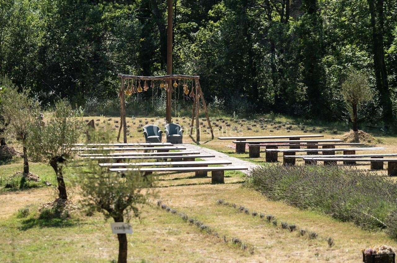 Chaises disposées sous une arche décorée, entourées de rangées de bancs en bois dans un cadre naturel verdoyant.