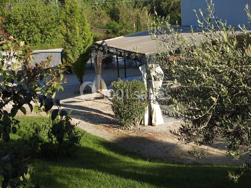 Vue d'un jardin avec une pergola ombragée, des arbustes, des arbres et un sol en gravier.