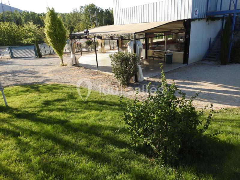 Terrasse couverte avec sol en béton, entourée de verdure et située près d'un bâtiment métallique.
