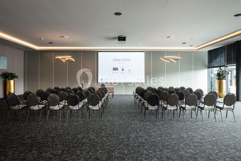 Location salle Nivelles (Brabant wallon) - Hôtel Nivelles Sud #6 Salle de conférence moderne avec rangées de chaises alignées face à un écran de projection allumé.