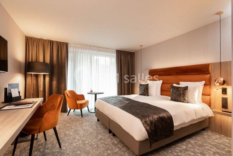 Location salle Nivelles (Brabant wallon) - Hôtel Nivelles Sud #11 Chambre d'hôtel moderne avec lit double, bureau, fauteuils orange, lampadaire et grande fenêtre avec rideaux.