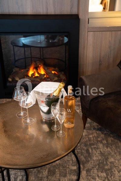Location salle Nivelles (Brabant wallon) - Hôtel Nivelles Sud #12 Bouteille de champagne dans un seau à glace, bouteille de vin, verres et cheminée allumée en arrière-plan.