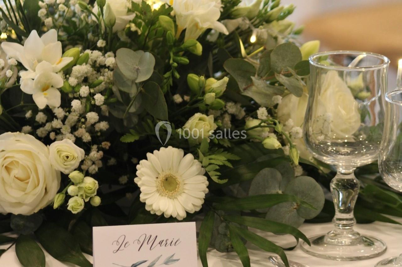 Centre de table floral avec roses blanches, gypsophiles, gerbera et feuillage, accompagné de verres et d'une carte ’Le Marié…