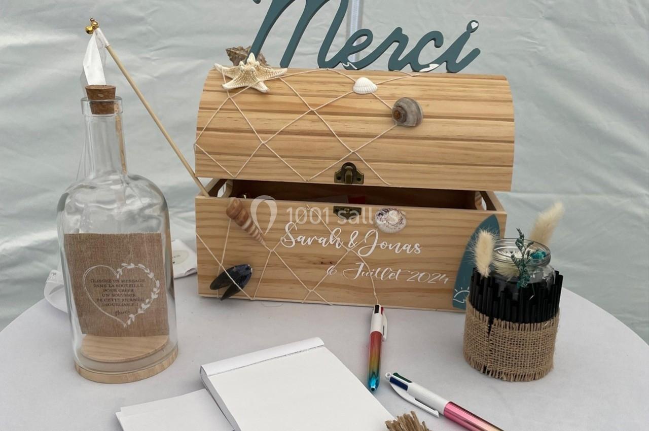 Coffre en bois décoré pour un mariage avec une inscription, entouré de stylos, papier et une bouteille en verre.
