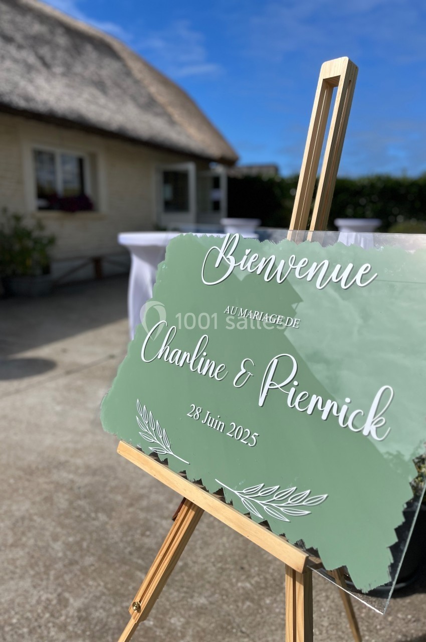 Panneau de bienvenue sur chevalet pour un mariage, devant une maison au toit de chaume et un ciel dégagé.