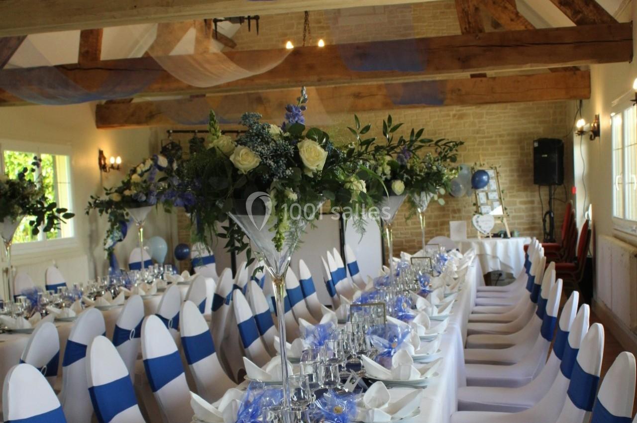 Salle de réception décorée pour un événement, avec des tables dressées, des chaises blanches ornées de rubans bleus et des…
